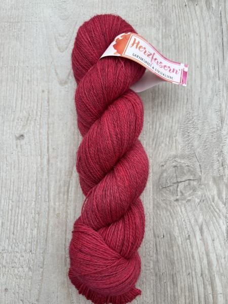 Ein Strang handgefärbter Wolle in einem warmen, tiefen Rotton liegt auf hellem Holz. Der Strang ist gedreht, die Melierung der Faser ist sichtbar, die Herzfasern-Banderole ist umgelegt.